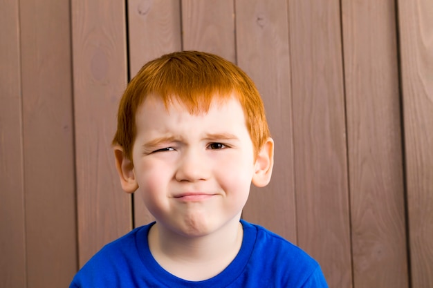 Un garçon aux cheveux roux plissant un œil sur un fond de planches marron à l'enfant sceptique