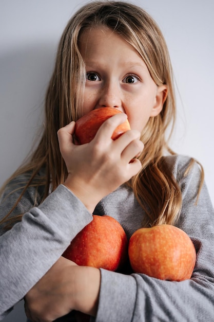 Garçon aux yeux écarquillés mordre une pomme tenant un tas dans son bras sur un mur blanc