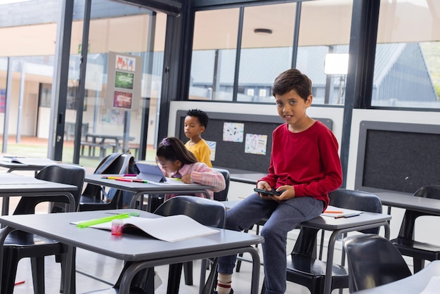 Un garçon biracial dans un pull rouge tient un téléphone intelligent dans une salle de classe avec un espace de copie