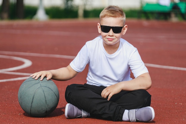 Garçon blond assis avec un ballon de basket sur le terrain de jeu. photo de haute qualité