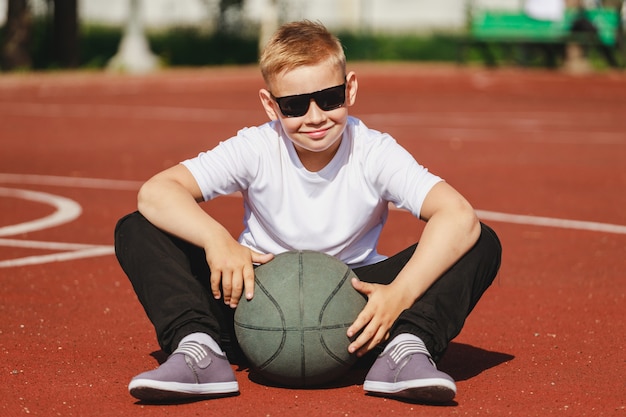 Garçon blond assis sur un terrain de basket avec ballon en été. photo de haute qualité