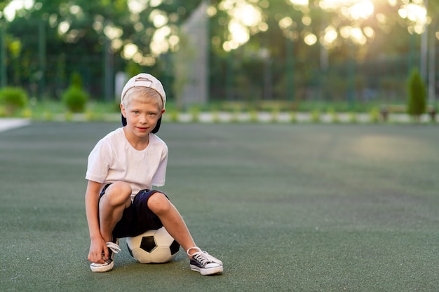 Garçon blond dans une casquette dans un uniforme de sport est assis sur un ballon de football sur le terrain de football