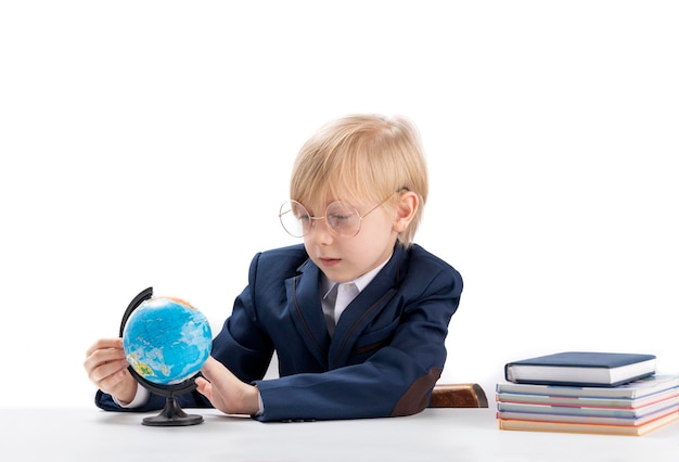 Garçon blond dans de grands verres et uniforme scolaire est assis au bureau et examine attentivement le globe Portrait d'écolier isolé sur fond blanc