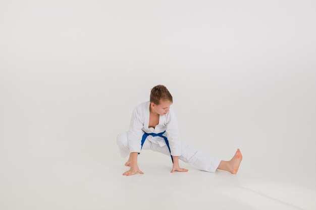 Garçon blond dans un kimono blanc avec une ceinture bleue fait un étirement sur un mur blanc