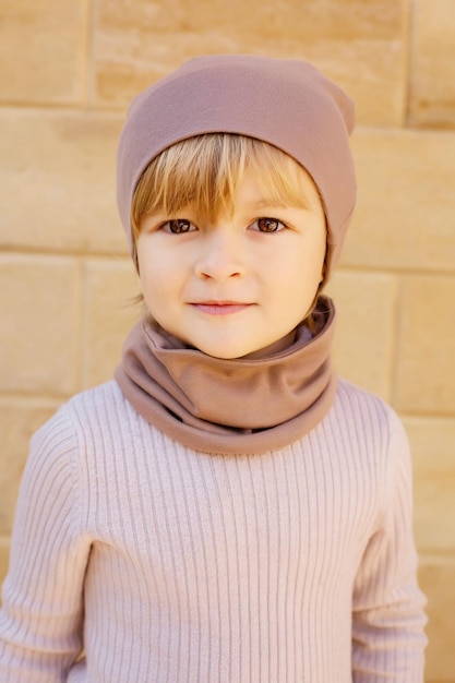 un garçon blond avec des fossettes sur ses joues dans un chapeau beige et un snood sur le fond d'un mur de pierre