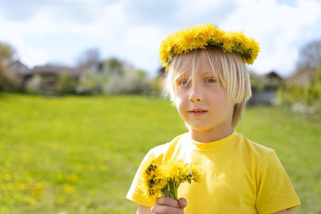 Photo garçon blond en guirlande de fleurs avec bouquet de pissenlits se tenir dans le pré les vacances d'été