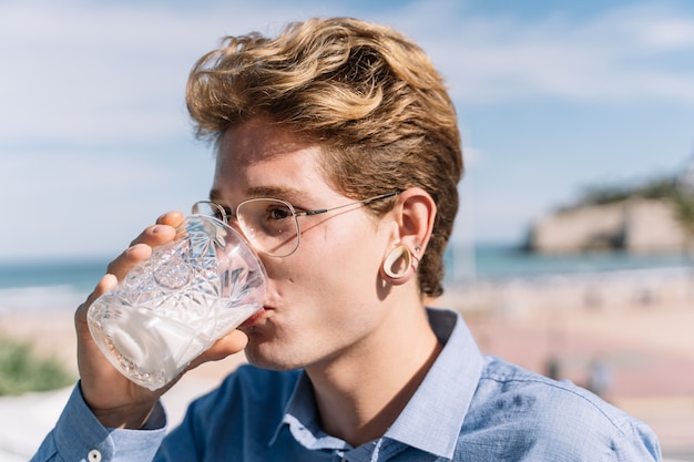 Garçon blond avec des lunettes et des piercings de l'eau potable dans un verre