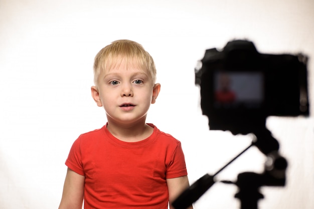 Un garçon blond sérieux enregistre une vidéo sur un appareil photo reflex numérique. Petit blogueur vidéo. mur blanc