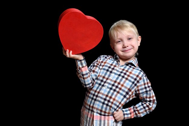 Garçon blond souriant en chemise à carreaux tenant une boîte en forme de coeur rouge.