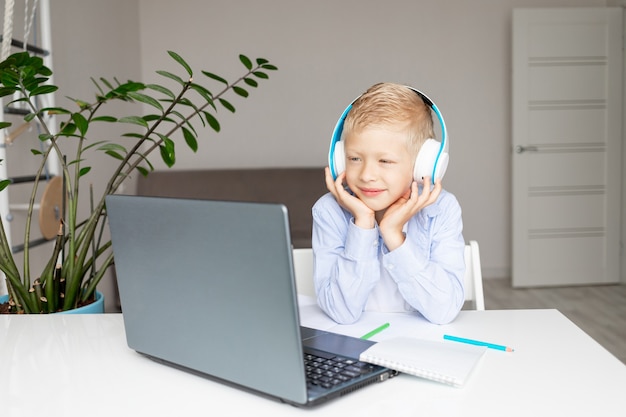 Un garçon blond souriant portant des écouteurs passe un appel vidéo via un ordinateur portable pendant l'apprentissage électronique à distance à la maison, concept de retour à l'école