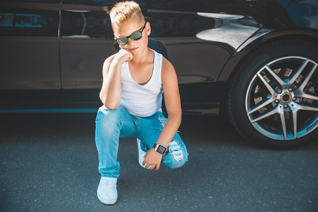 Garçon blond en t-shirt blanc et jeans et lunettes de soleil à côté de la voiture