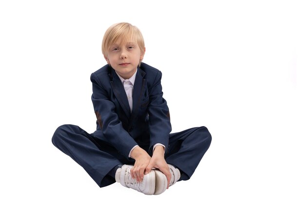 Garçon blond en uniforme scolaire est assis avec ses mains sur des baskets Portrait d'élève de 1ère ou 2ème année isolé sur fond blanc