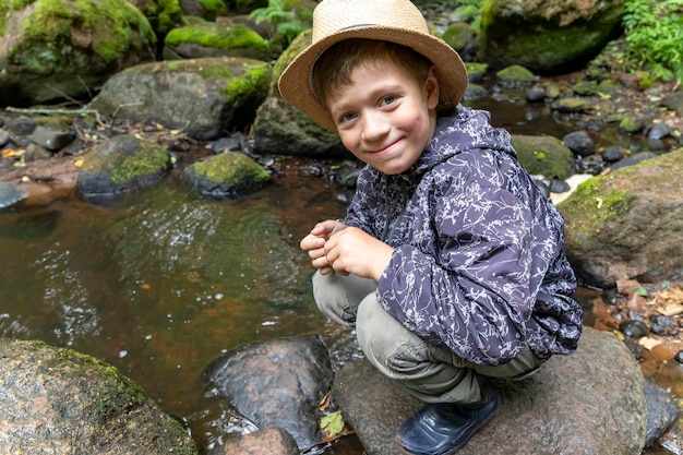 Un garçon en bottes de caoutchouc, un chapeau de voyageur est accroupi près d'un ruisseau forestier