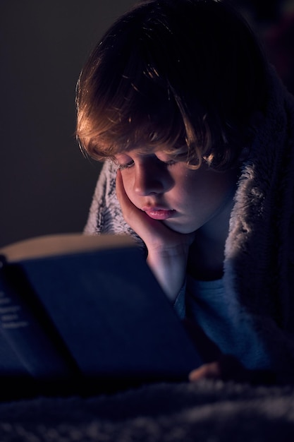 Garçon calme enveloppé dans une couverture chaude tout en lisant un livre intéressant sur le lit dans une pièce sombre avec une faible lumière à la maison