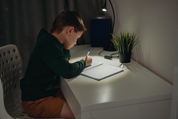 Garçon caucasien faisant ses devoirs à la maison dans une pièce sombre à la lumière de la lampe