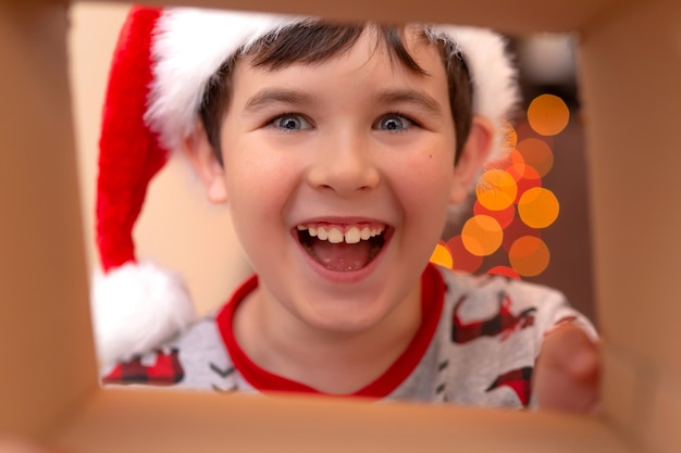 Un garçon caucasien heureux et beau dans un pull du Nouvel An et un chapeau de père Noël s'écrase dans une boîte