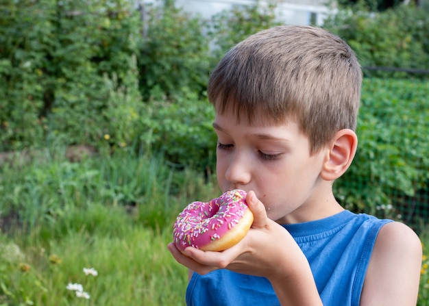 Un garçon caucasien mange un beignet rose. enfant tient une friandise dans le parc.