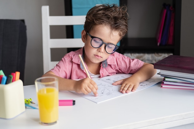 Un garçon caucasien mignon et sérieux à lunettes étudie à la maison, fait ses devoirs. L'écriture. Ordinateur portable sur le bureau. concept d'éducation à distance. Quarantaine. Retour au concept de l'école.