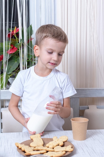 Un garçon caucasien mignon verse du lait dans un gobelet en papier artisanal sur la table à côté de biscuits pour le Père Noël. Concept de la veille de Noël. L'enfant prépare un festin pour Santa