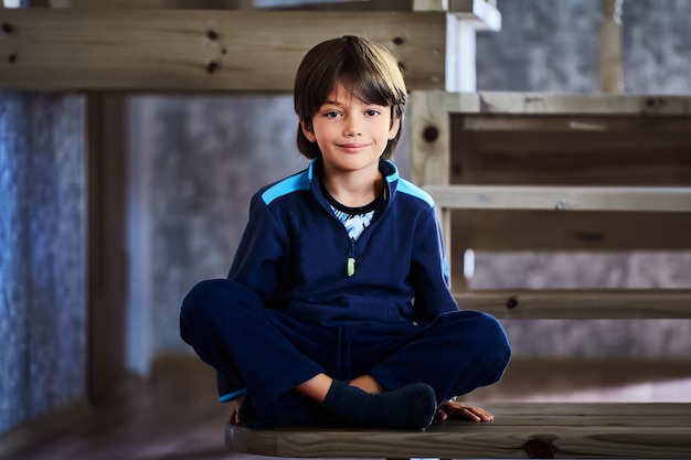 Un garçon caucasien de sept ans est assis les jambes croisées sur l'escalier en bois de sa maison.