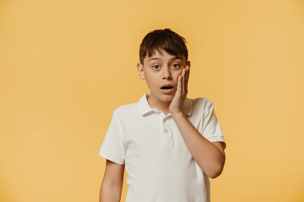 Photo un garçon caucasien stupéfait choqué en t-shirt blanc garde la mâchoire baissée, touche la joue avec la main, halète d'excitation, regarde avec des yeux embués, isolé sur le mur jaune, a une expression désespérée.