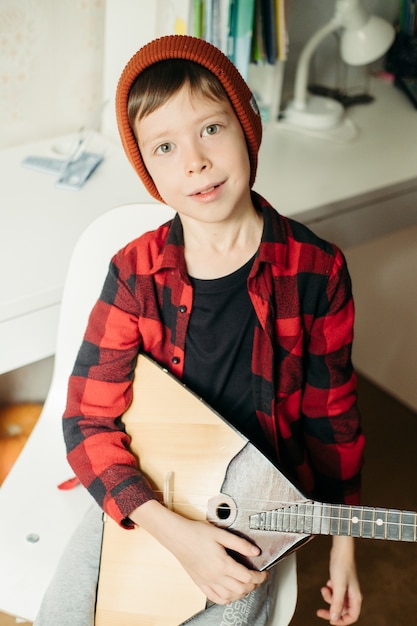 Un garçon avec un chapeau rouge et une chemise à carreaux joue de la balalaïka. Beau garçon tenant sa guitare. Cours de musique à domicile. Hobby pour l'âme. enseignement de la musique à domicile