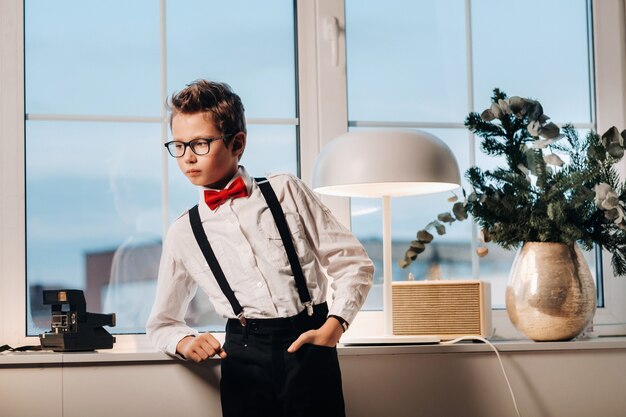 Un garçon en chemise avec un nœud papillon rouge et des lunettes se tient à la fenêtre.