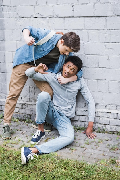 Garçon en colère en chemise bulling va frapper un garçon afro-américain effrayé en sweat à capuche