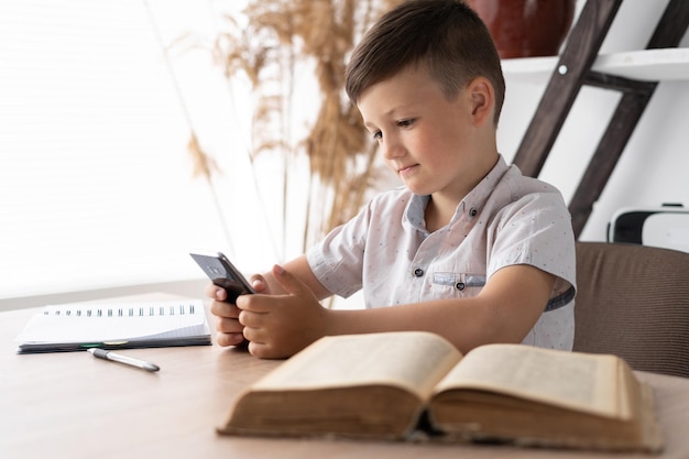 Garçon concentré jouant à un jeu sur le téléphone portable tout en étant assis à la table fait ses devoirs Étudiant accro aux gadgets Concept pour les jeux en ligne ou les réseaux sociaux