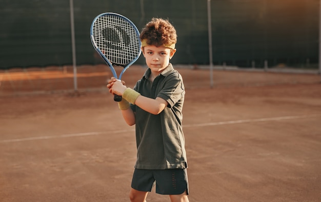 Garçon confiant en uniforme avec une raquette dans les mains regardant la caméra tout en jouant au tennis sur le court pendant l'entraînement d'été