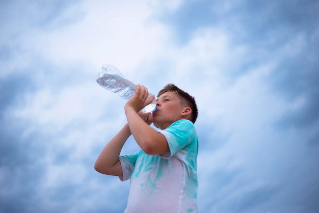Un garçon contre le ciel boit de l'eau d'une bouteille en plastique soif chaleur ciel nuageux