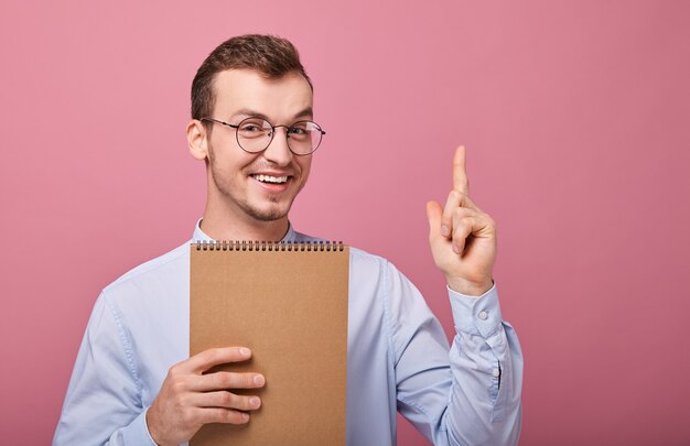 Garçon cool assez souriant dans une chemise doucement bleue tient un cahier à feuilles mobiles marron et apparaît