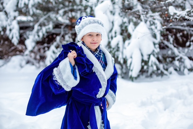 Un garçon en costume de père Frost avec un sac de cadeaux contre les arbres enneigés. Jour d'hiver