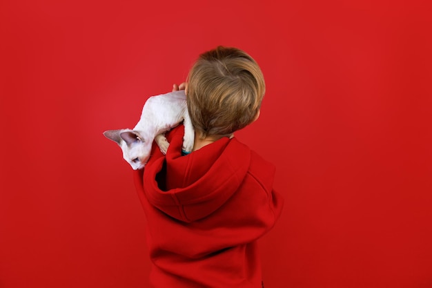 Un garçon en costume rouge se dresse sur un fond rouge tourné le dos tient un petit chaton blanc sur son épaule qui regarde autour de lui