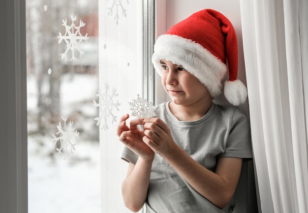 Un garçon dans un bonnet de Noel tient un flocon de neige dans ses mains et regarde par la fenêtre