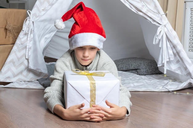 Un garçon dans un bonnet de Noel tient sa grande boîte-cadeau pour le Nouvel An de Noël allongé dans un wigwam de tente pour enfants dans la pépinière