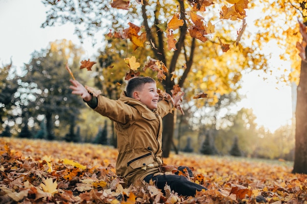 Garçon dans une boucle jaune jette des feuilles d'automne jaunes dans le parc en automne