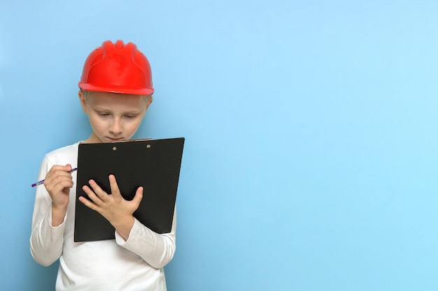 Garçon dans un casque de construction protecteur se concentrant sur l'écriture de quelque chose avec un stylo sur une tablette sur fond bleu avec espace de copie