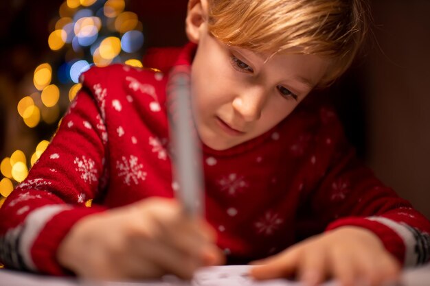 Un garçon dans un chandail de Noël rouge sur le fond d'un arbre de Noël dessine avec diligence derrière une lampe qui brille sur lui