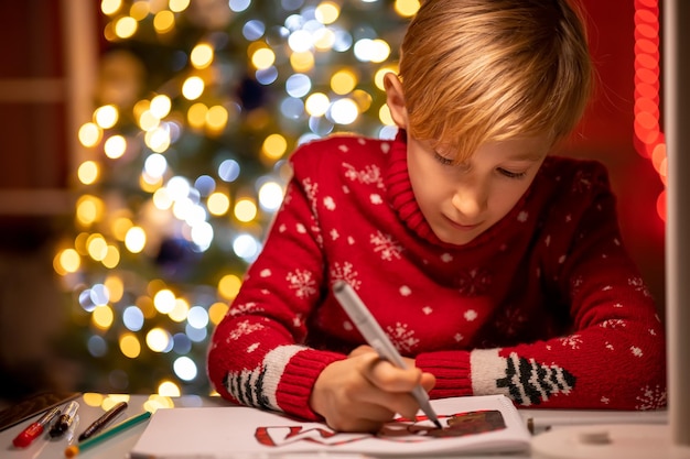 Un garçon dans un chandail de Noël rouge sur le fond d'un arbre de Noël se pencha pour faire un dessin avec un marqueur