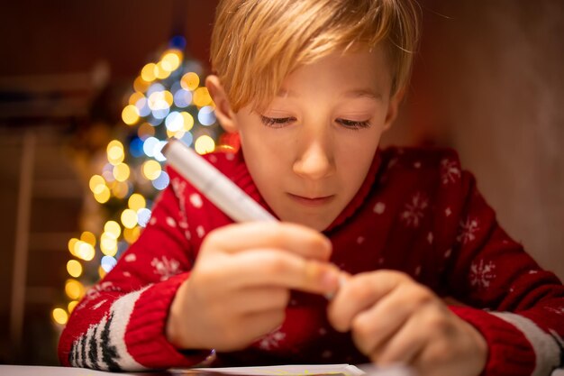 Un garçon dans un chandail de Noël rouge sur le fond d'un sapin de Noël est en train de dessiner dans sa chambre sous une lampe