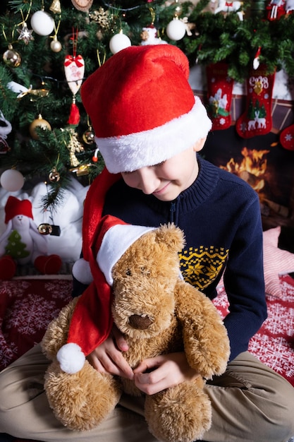 Un garçon dans un chapeau de Père Noël sourit à un jouet ours dans le même chapeau