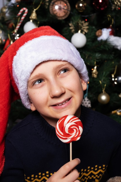 Un garçon dans un chapeau de Père Noël tient du caramel sur un bâton et regarde au loin des rêves de cadeaux et de bonbons pour les vacances