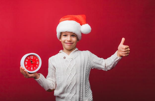 Un garçon dans un chapeau de Père Noël tient une montre sur un fond de studio rouge le concept de Noël et du nouvel an un enfant en vêtements tricotés