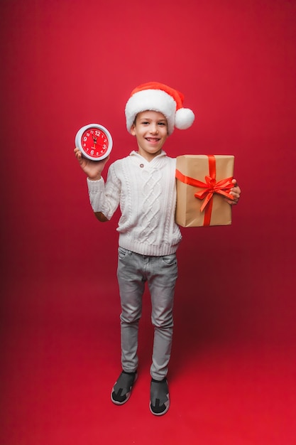 Un garçon dans un chapeau de Père Noël tient une montre sur un fond de studio rouge le concept de Noël et du nouvel an un enfant en vêtements tricotés