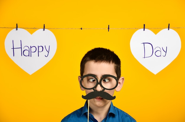 Un garçon dans une chemise bleue sur fond jaune avec un cœur et une inscription un jour de fête est tordu et habille des lunettes et une moustache