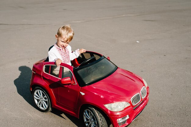 Le garçon dans une chemise brodée roule sur une magnifique voiture rouge sur la route Close up Little baby boy Le bébé conduit la voiture