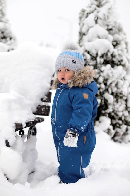 Un garçon dans une combinaison bleue et un chapeau voit la neige pour la première fois et se tient près d'un banc dans le contexte d'un arbre de Noël enneigé dans le parc.