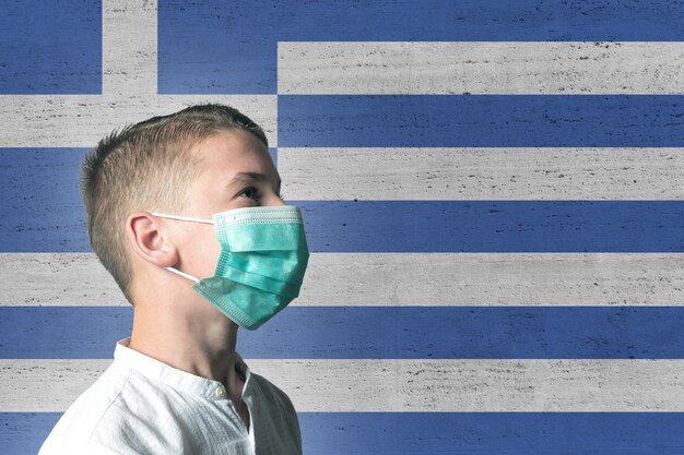 Garçon dans un masque médical sur son visage sur fond de drapeau de la Grèce.