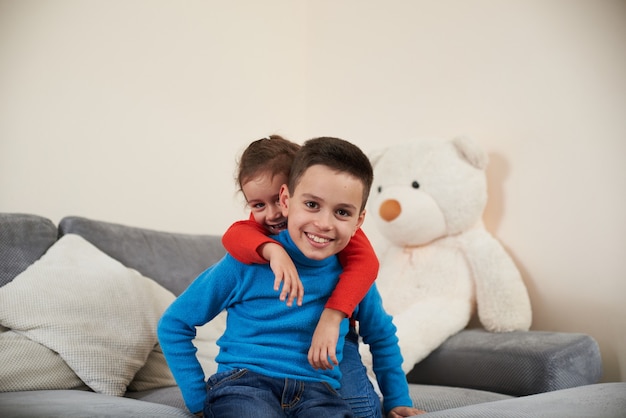 Un garçon dans un pull bleu est assis sur le canapé tandis que sa sœur le serrant derrière son dos et souriant à la caméra